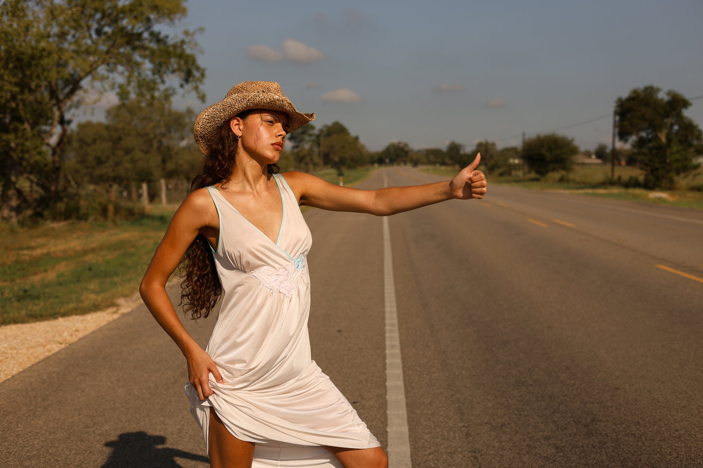 1970s white maxi slip dress with floral appliqué and lace trim styled western, model hitchhiking on sunlit country road