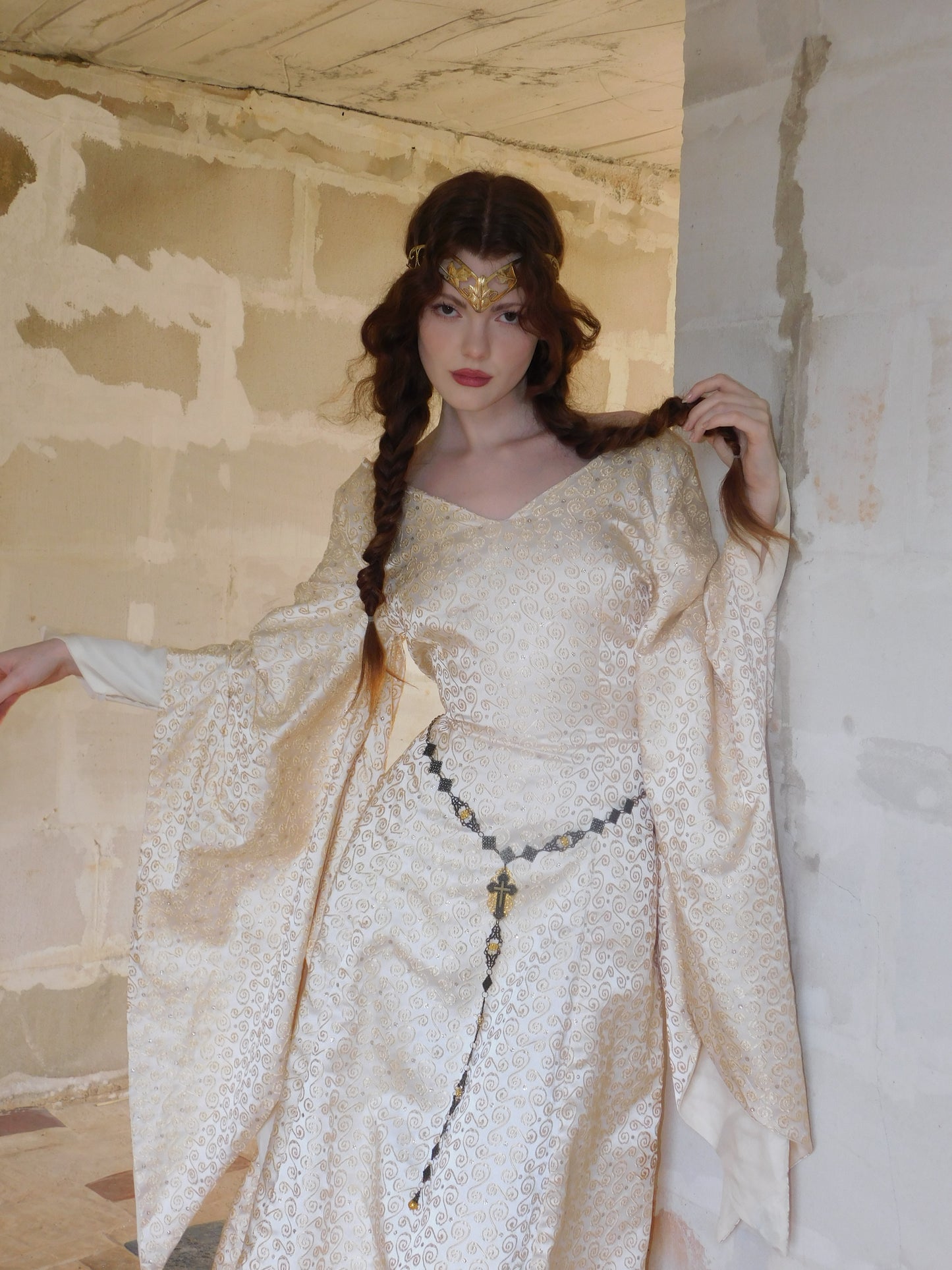 Model wearing a 1930s Spanish silk gown with metallic gold swirl embroidery, standing in a softly lit hallway. Her hair is styled in two long braids with a golden crown headpiece, evoking an elven or medieval fantasy look.