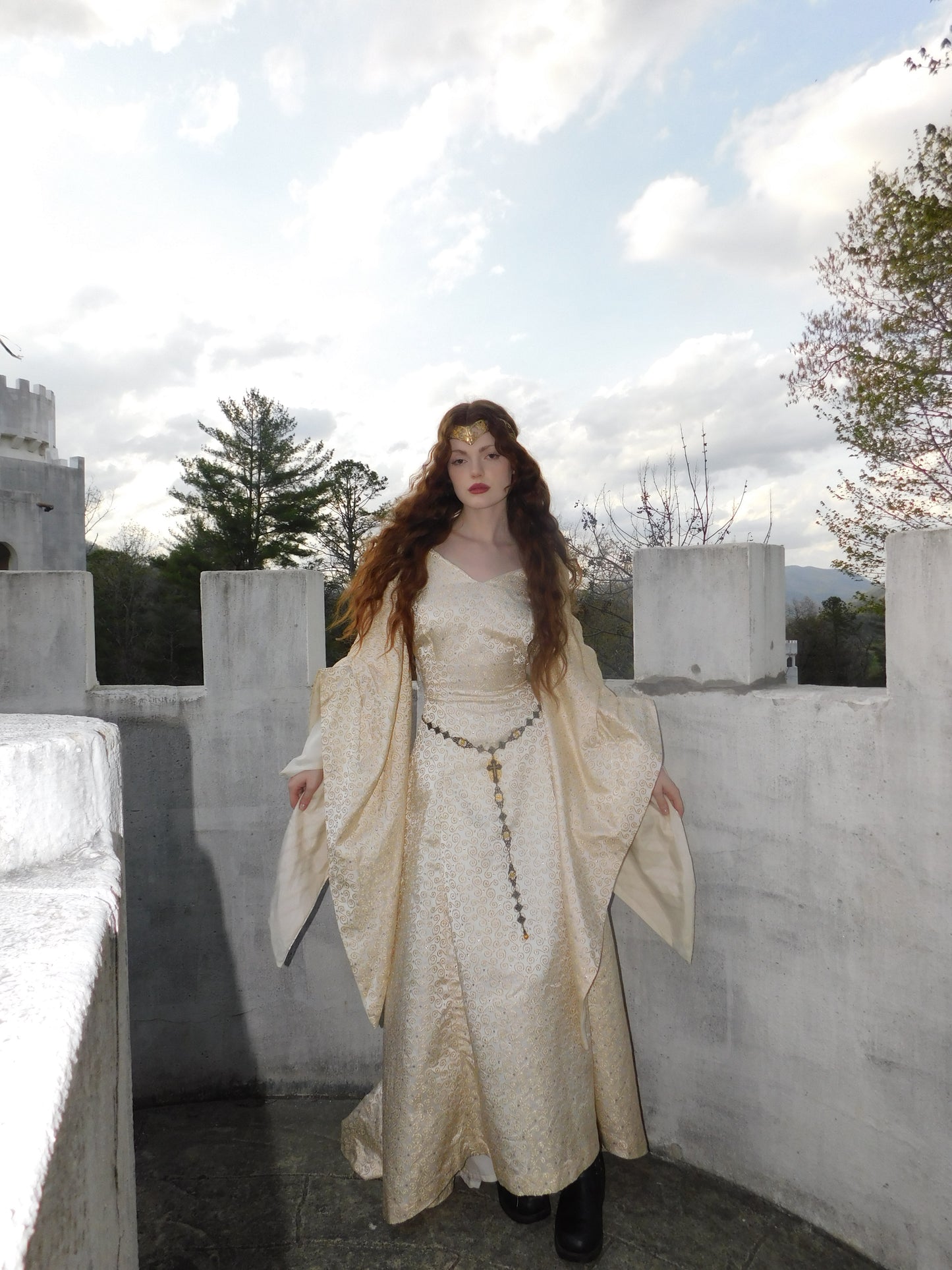 Windblown model in medieval-style silk dress stands on castle balcony with clouds behind.