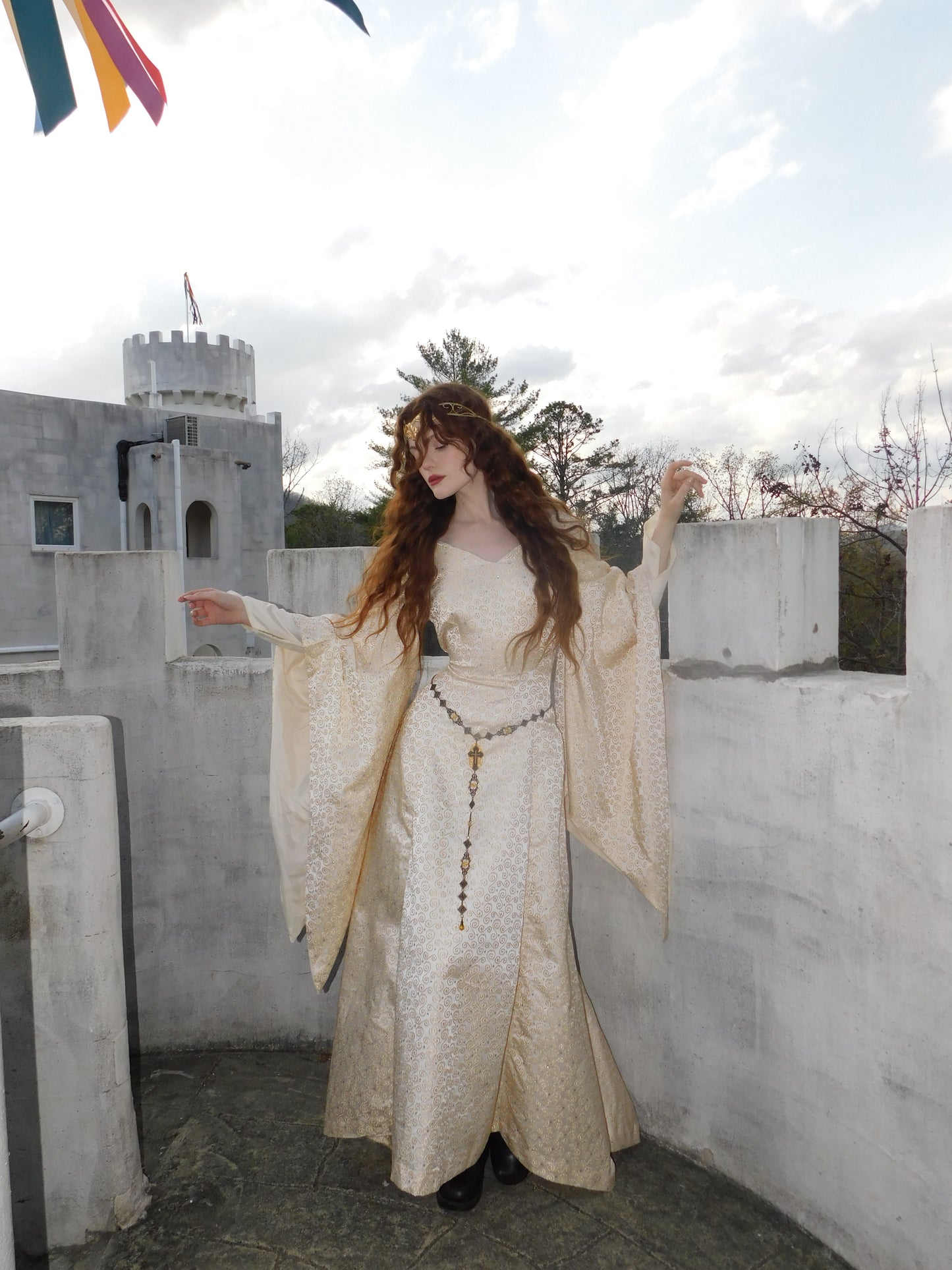 Full-body image of model in embroidered 1930s medieval-style gown with trumpet sleeves and vintage crown, posing on a castle rooftop under festival flags.