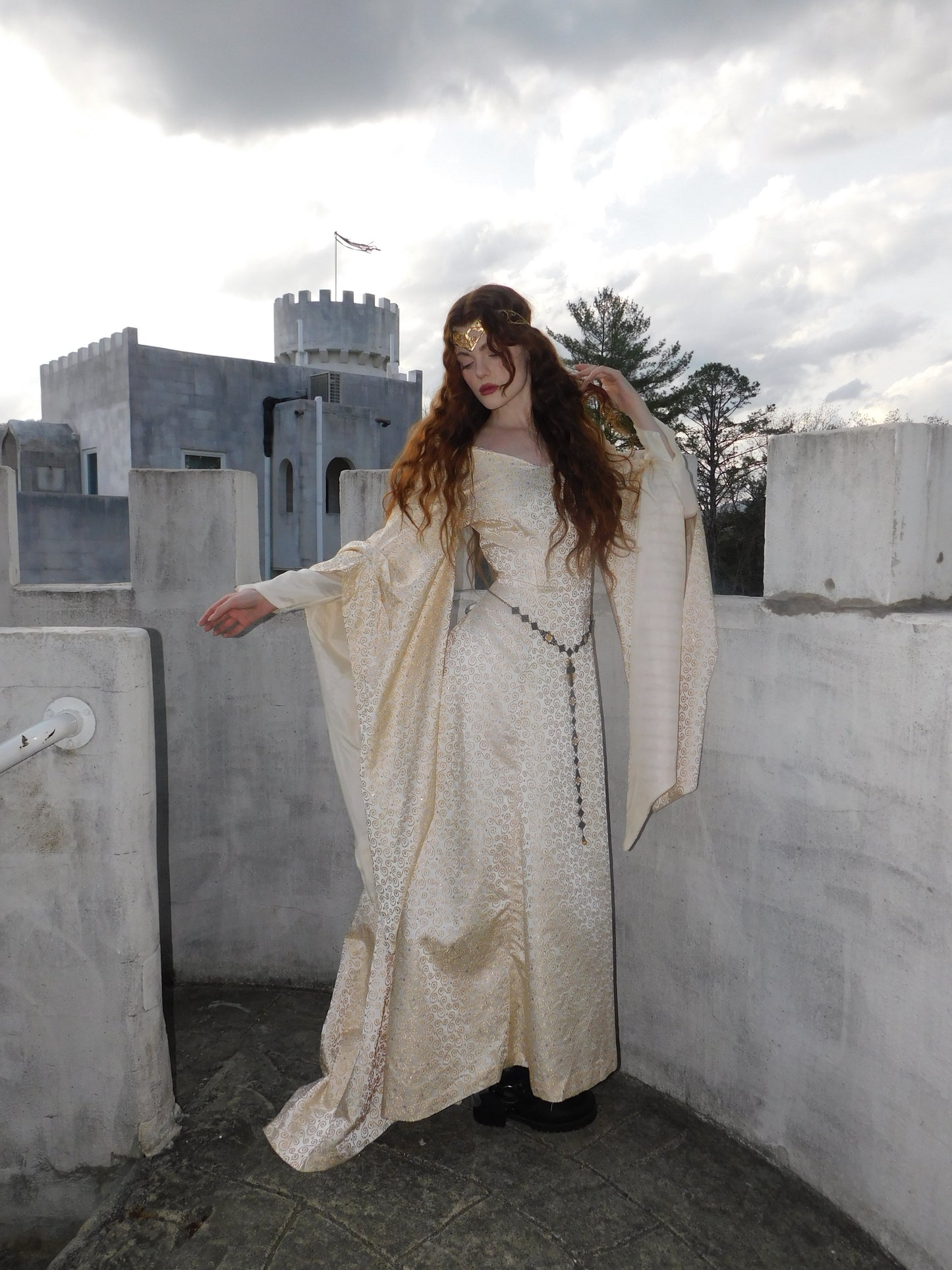 Model wearing medieval-style 1930s vintage gown with long flared sleeves and flowing silhouette, standing on castle balcony at dusk.