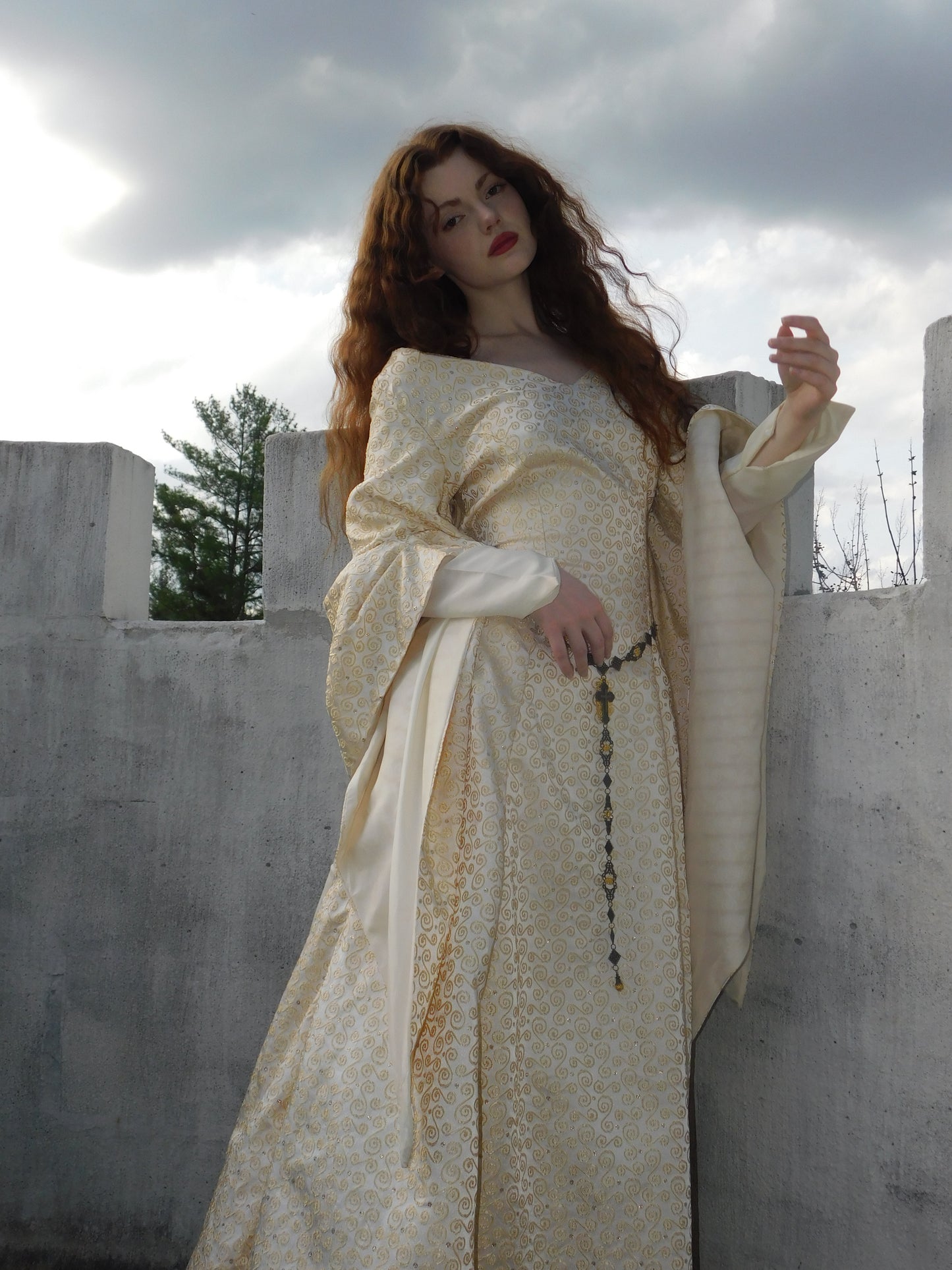 Model in a 1930s vintage Spanish silk gown with gold embroidery, long medieval sleeves, and rosary belt, standing against a cloudy sky on a stone terrace.