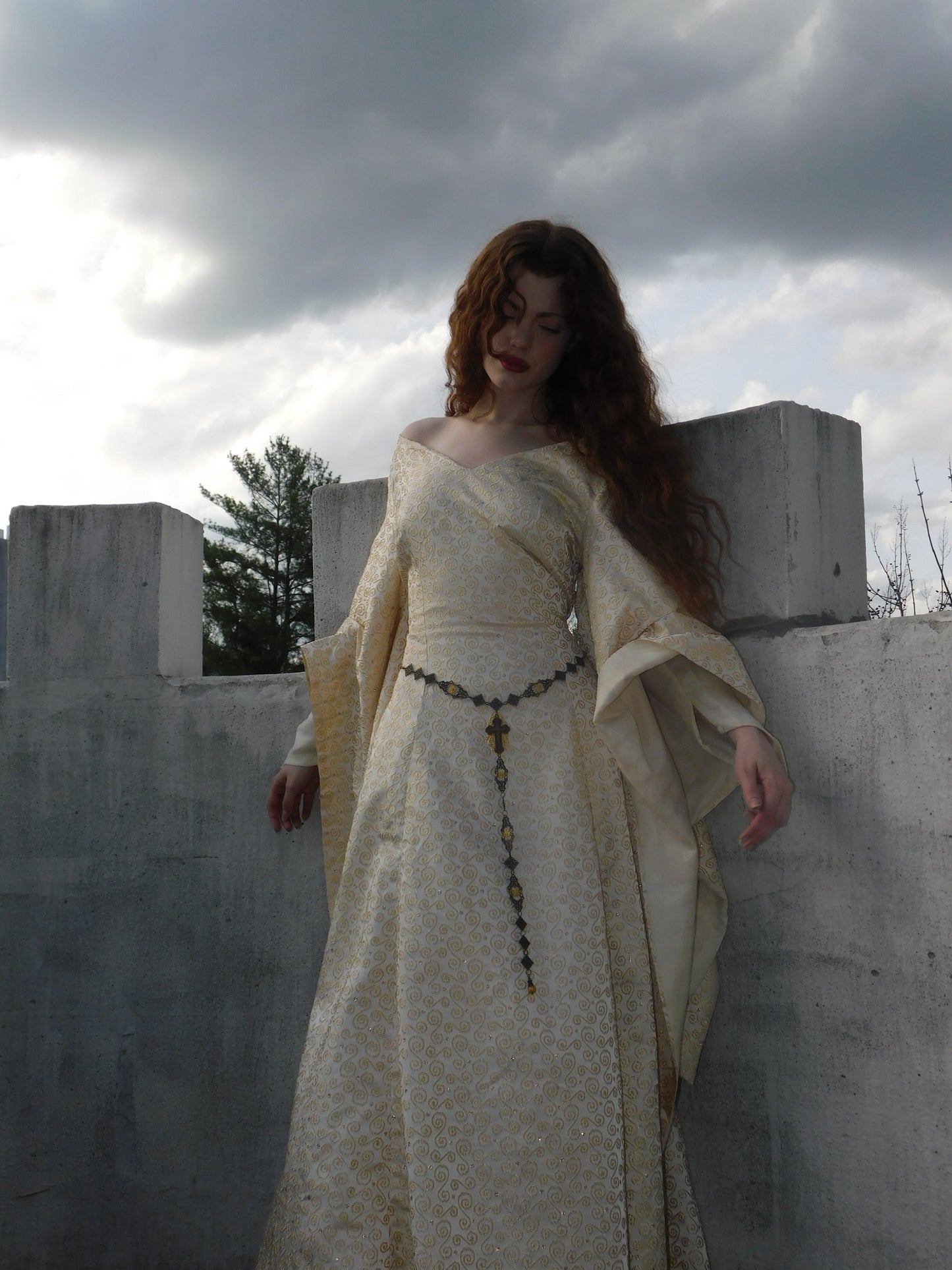 Model in a 1930s Spanish silk embroidered gown gazing downward with soft curls and flowing sleeves, set against castle stonework and cloudy sky.