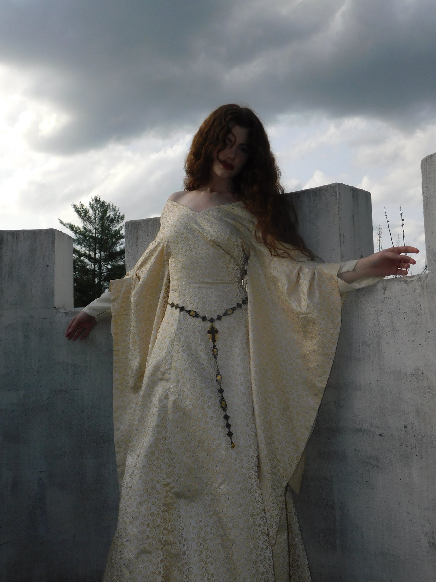 1930s vintage gown with gold embroidery and medieval sleeves shown with arms outstretched, framed by castle battlements and stormy light.