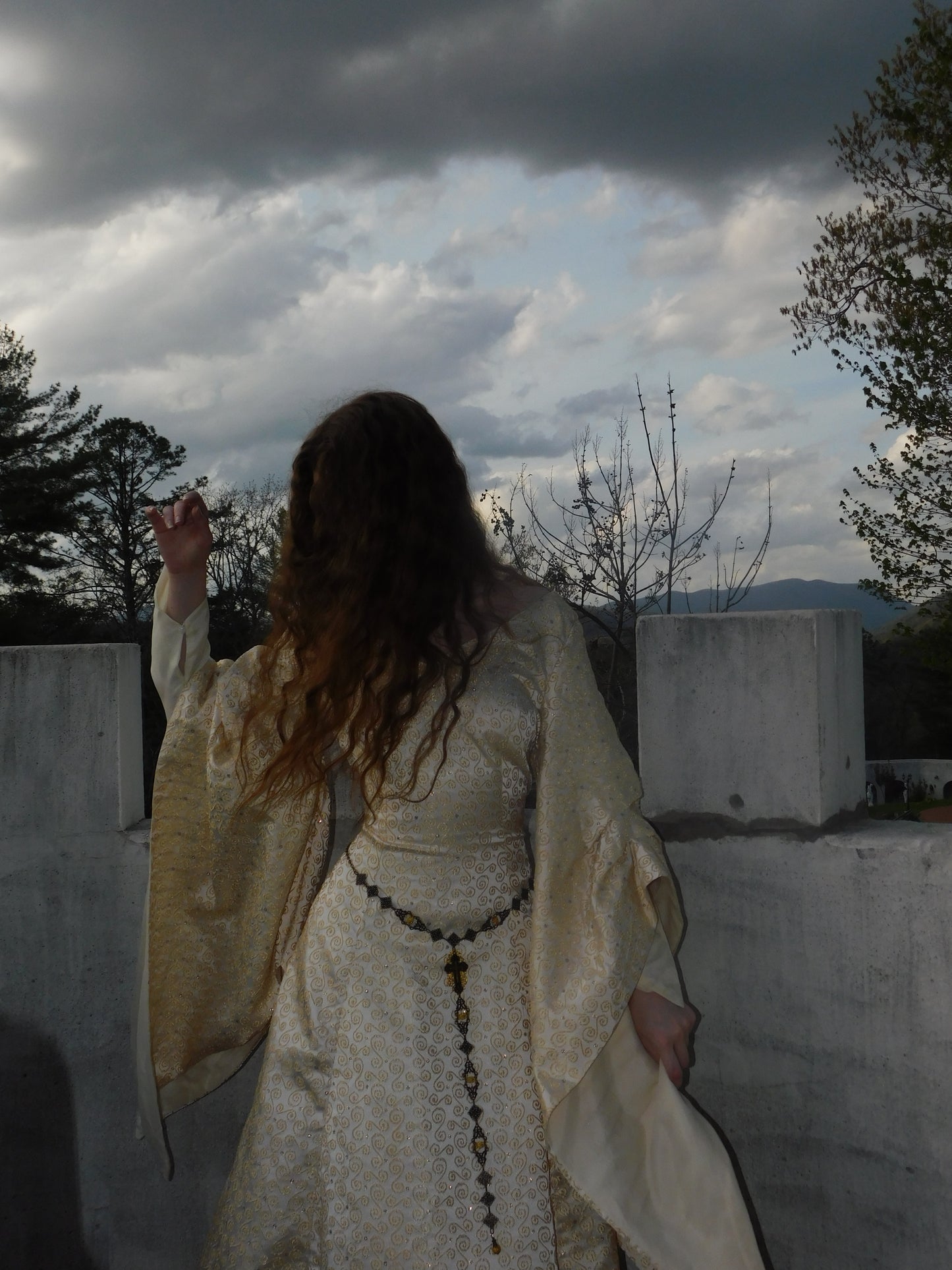 Model in gold brocade 1930s gown walking away with arm raised, medieval flared sleeves shown from behind, castle rooftop in background.