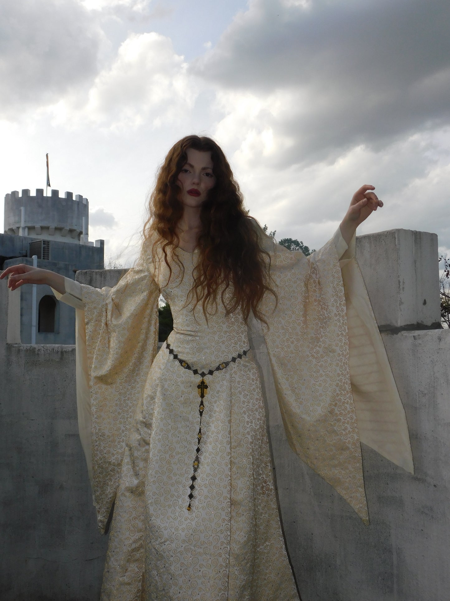 Model in a gold embroidered 1930s silk gown with medieval sleeves, arms lifted slightly, standing before a castle turret under dramatic skies.