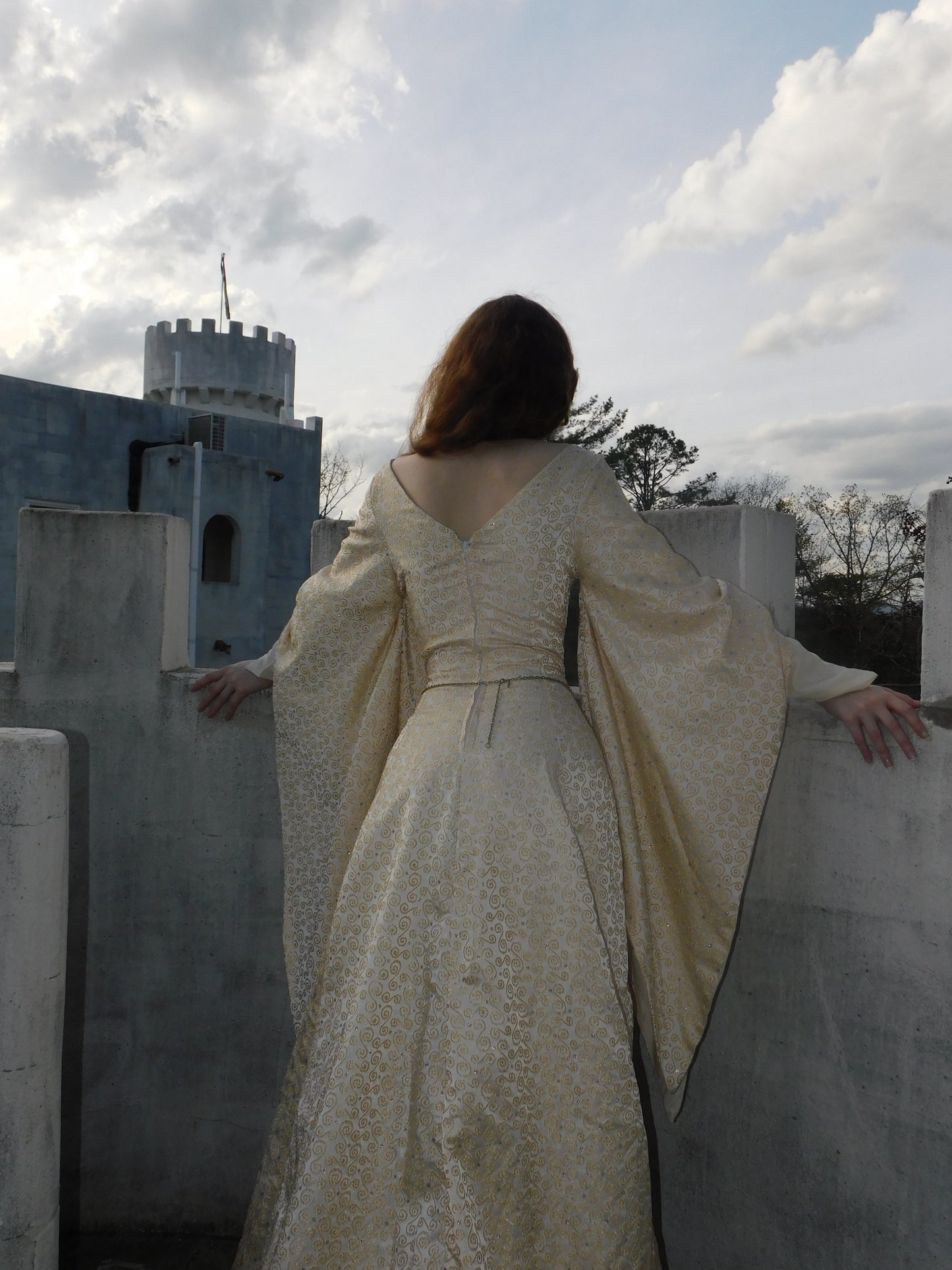 Full back view of medieval-style gown with intricate embroidery and draped sleeves, model’s arms resting on castle ledge under pale sky.