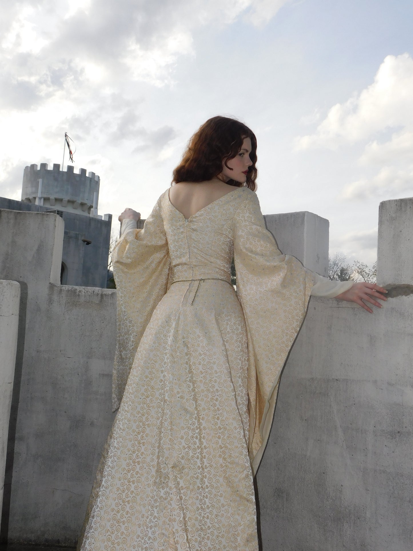 Side profile of model in embroidered vintage gown with long flared sleeves, sunlight catching curls as she looks back.