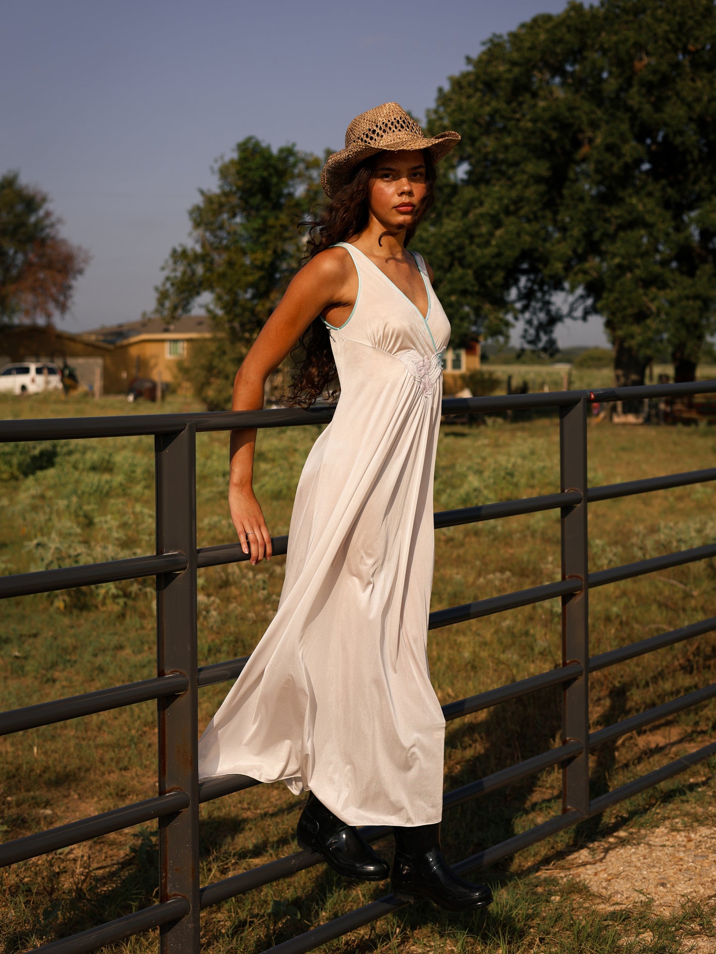 Model in 1970s white maxi slip dress with pastel floral appliqué leaning on ranch fence, styled with cowboy boots and hat
