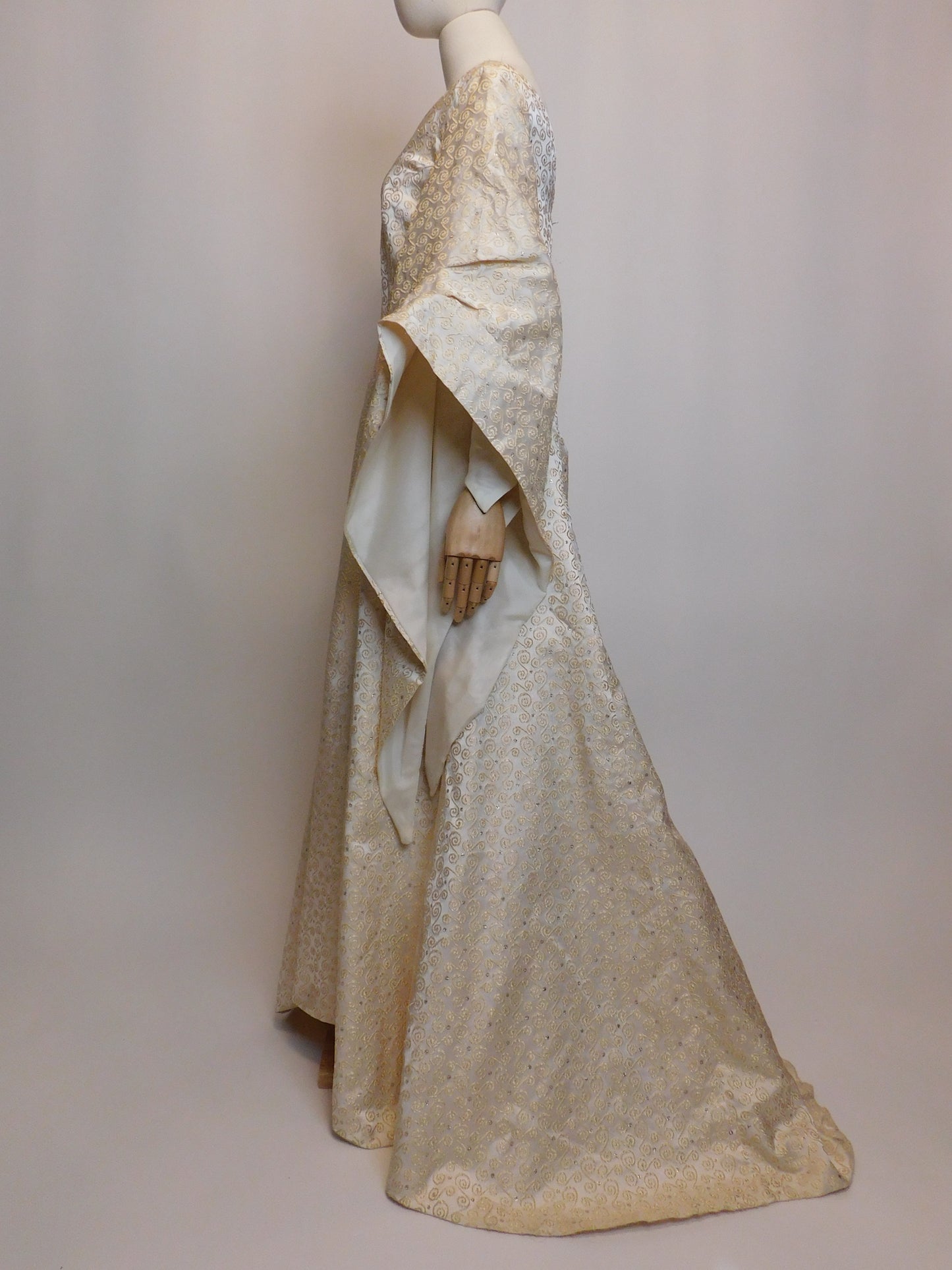Side profile of a 1930s Spanish silk gown displayed on a mannequin, featuring a floor-length silhouette and exaggerated medieval-inspired sleeves. The ivory silk fabric is embroidered with intricate golden spiral motifs.