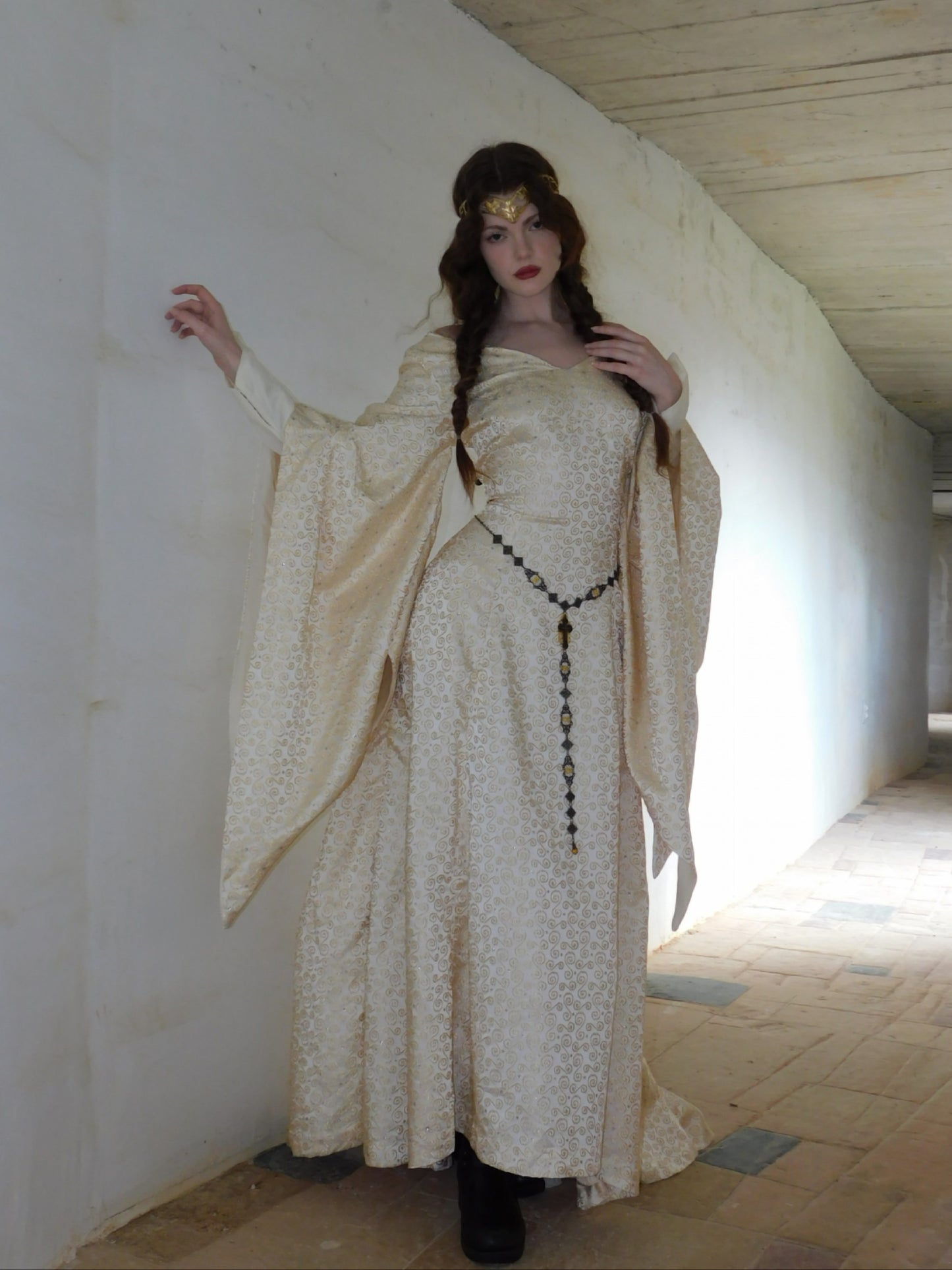 Full-length view of model in 1930s Spanish silk gown walking down a castle corridor. The dress features wide flowing sleeves and a rosary-style belt. Her posture and styling give a regal, ethereal presence.