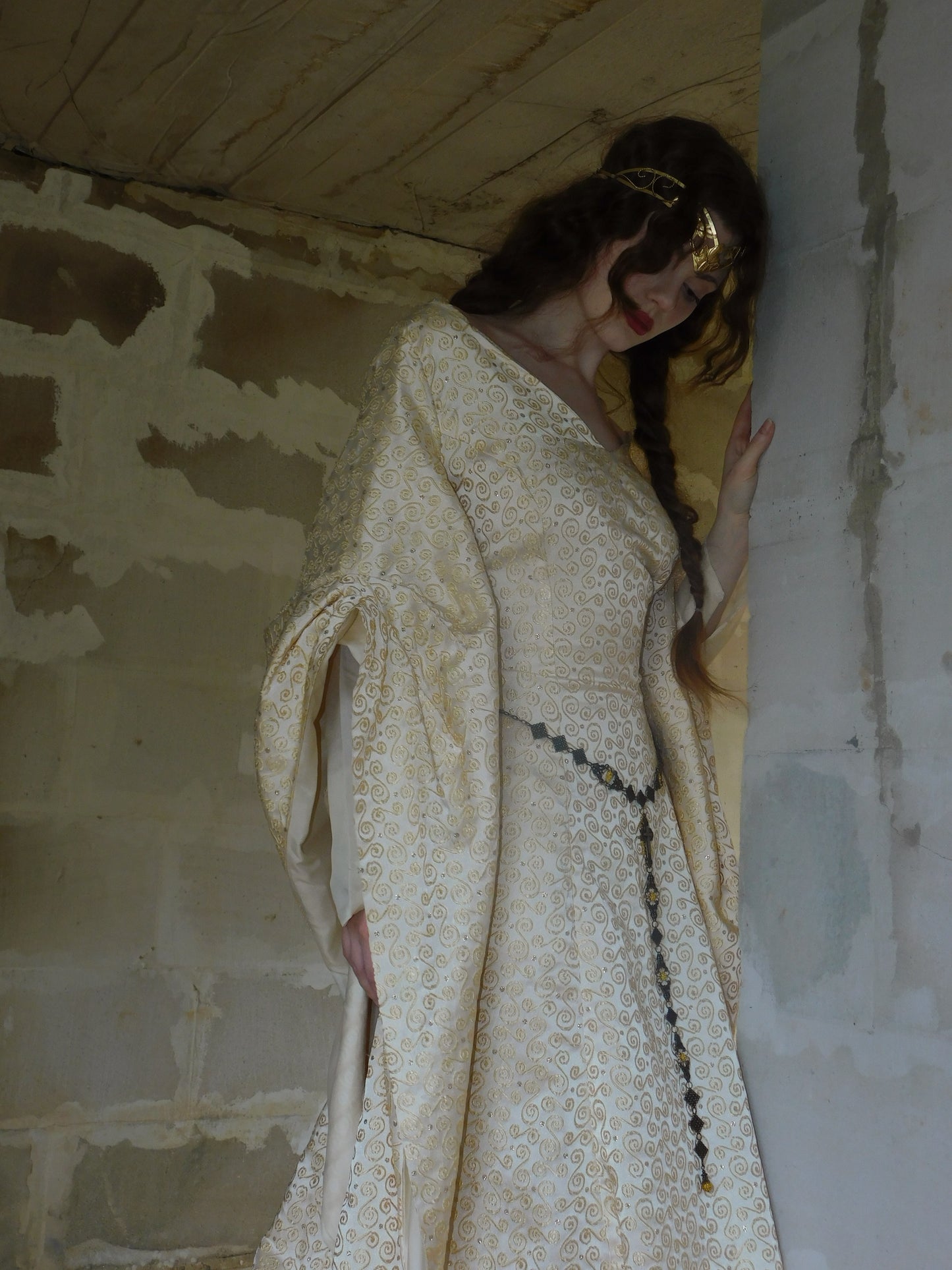 Woman in 1930s Spanish silk gown and gold crown standing in a stone alcove, looking down pensively.