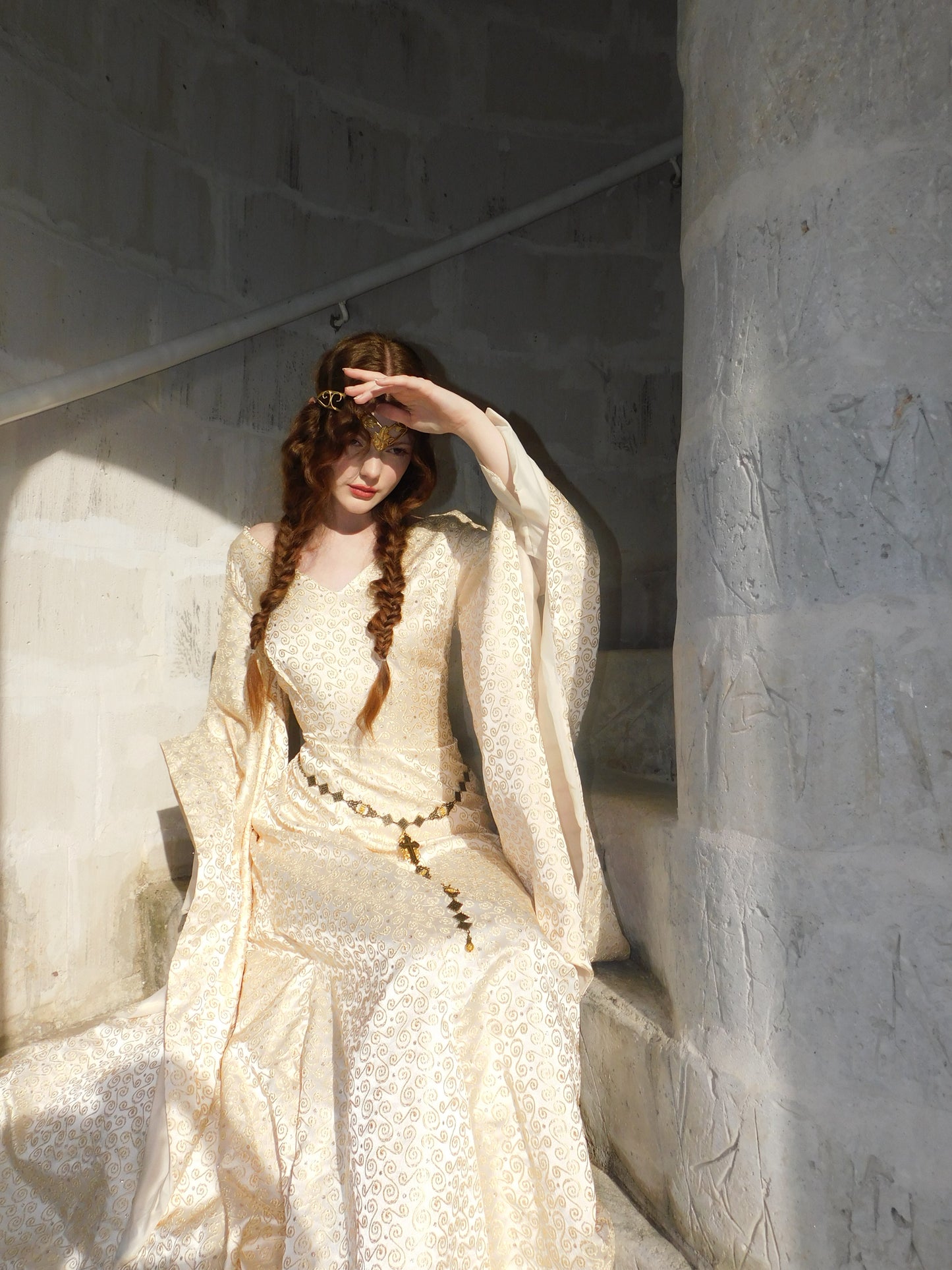 Model in embroidered gown sits in sunlit stone alcove, smiling with hand raised to shade eyes.