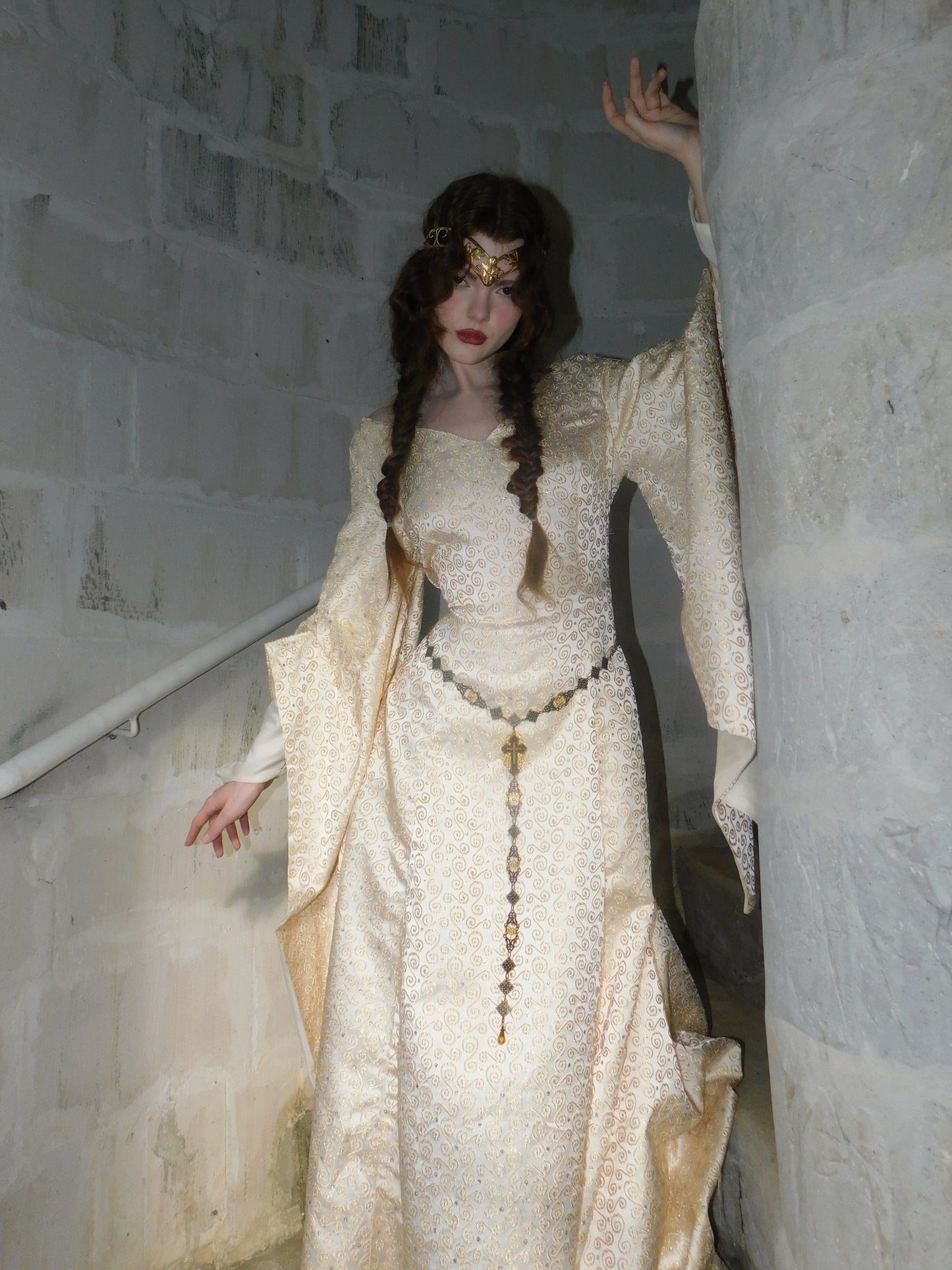 Model wearing a 1930s Spanish silk medieval-style gown with gold embroidery, standing in a stone stairwell with hand gracefully raised.
