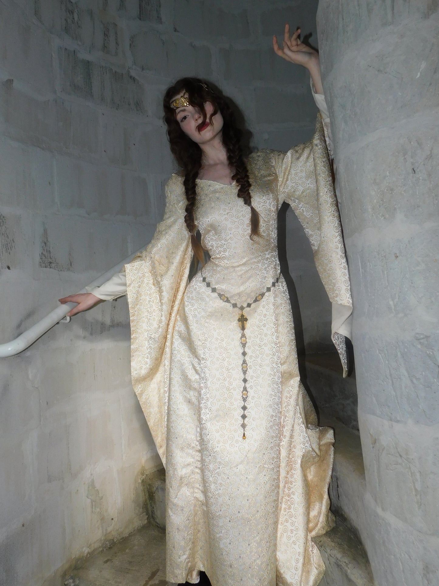 Model wearing 1930s vintage Spanish silk gown with gold embroidery, styled with a medieval circlet, raises hand in stone stairwell.