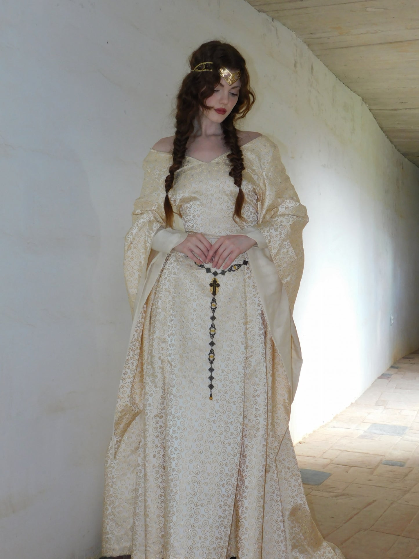 Model stands with hands folded, gazing down in a cream-gold 1930s Spanish silk gown with ornate swirl motifs. The softly illuminated corridor behind her enhances the romantic, historical mood of the image.