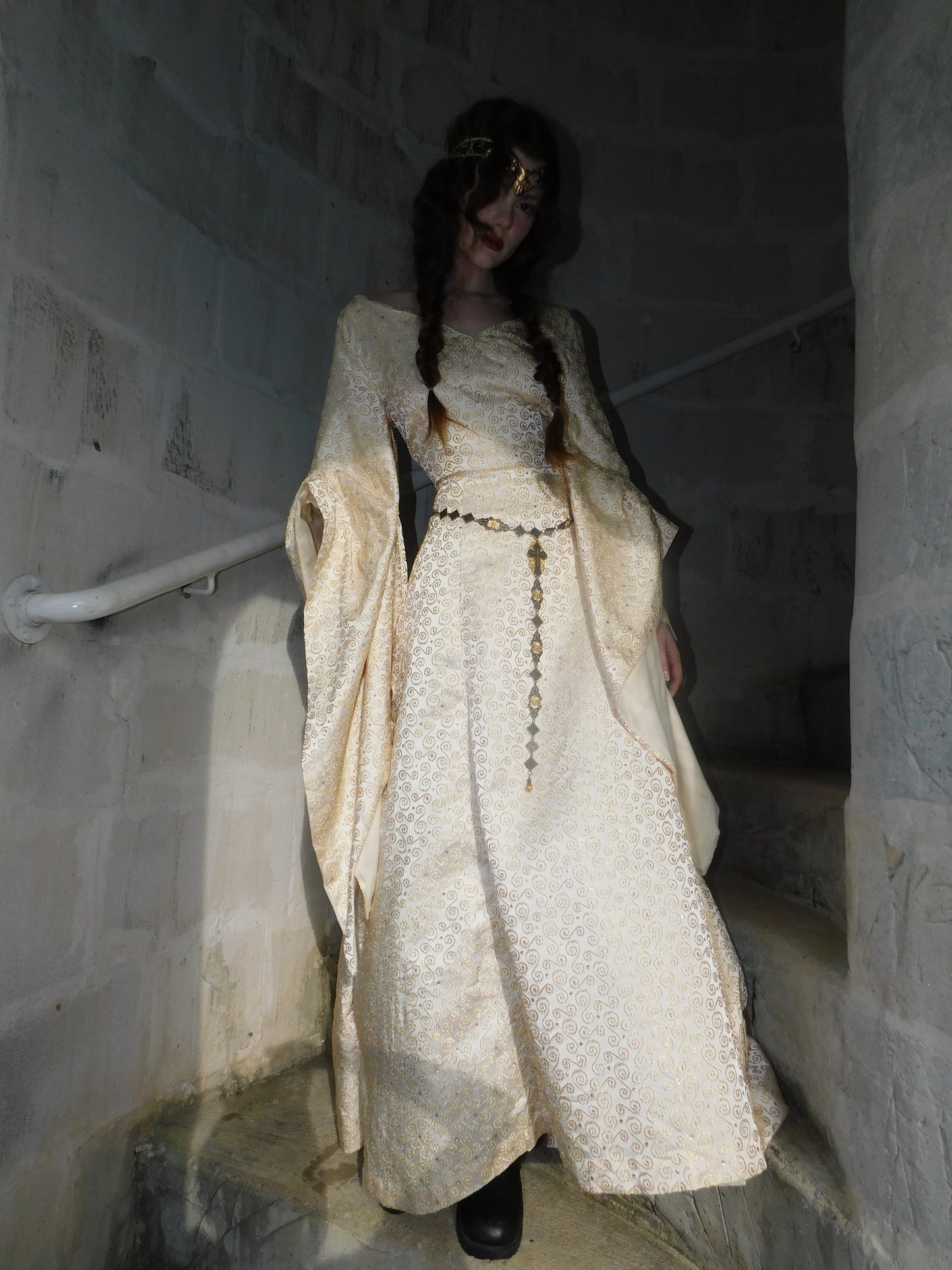 Model in vintage 1930s gold embroidered gown gazes downward, framed by a stone spiral staircase, showing full gown silhouette and rosary belt.