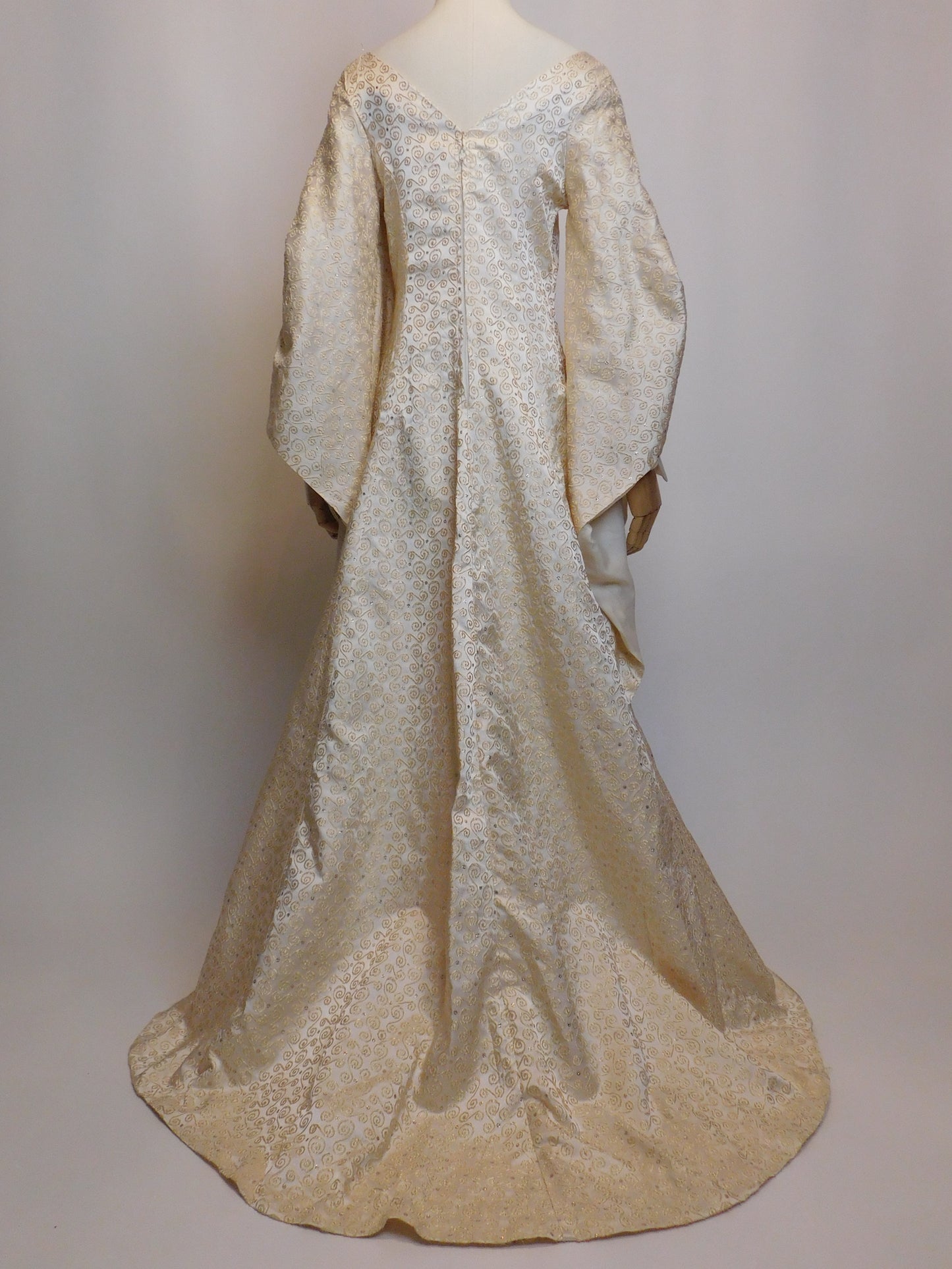 Full-length back view of a 1930s Spanish silk gown displayed on a mannequin. Features a dramatic train, V-shaped back neckline, and flowing brocade fabric with metallic gold embroidery.