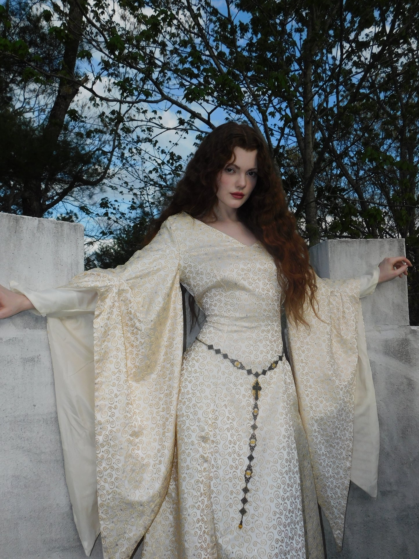 Woman in a 1930s Spanish silk gown with dramatic sleeves and intricate gold patterning, standing in front of a stone wall with a forest in the background.
