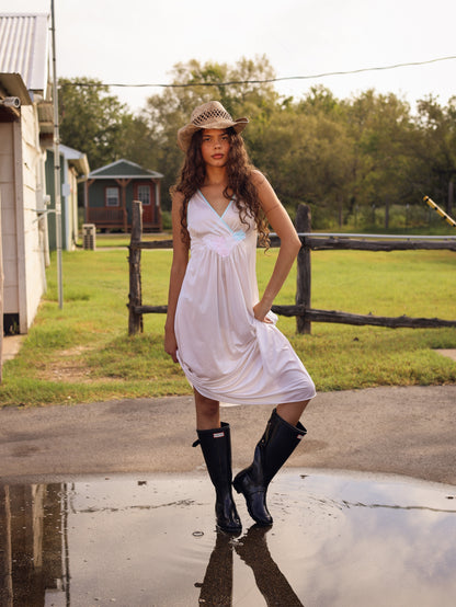 Model wearing 1970s white maxi slip dress with pastel floral appliqué and cowboy hat, standing on wet pavement after rain
