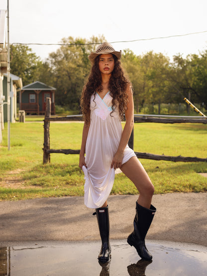 Model in 1970s white maxi slip dress with pastel floral appliqué posing in rural setting after rainfall, modern prairie look