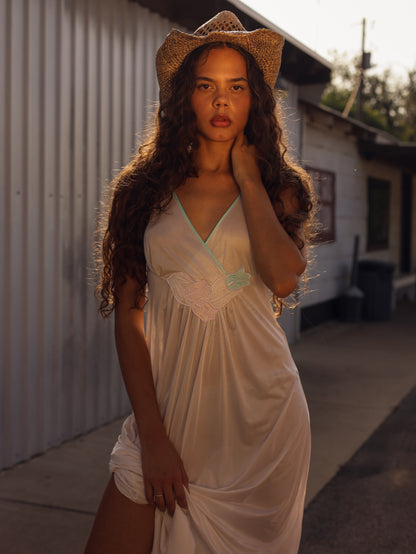 Close-up of model in 1970s white maxi slip dress with pastel floral appliqué, glowing in warm golden-hour sunlight