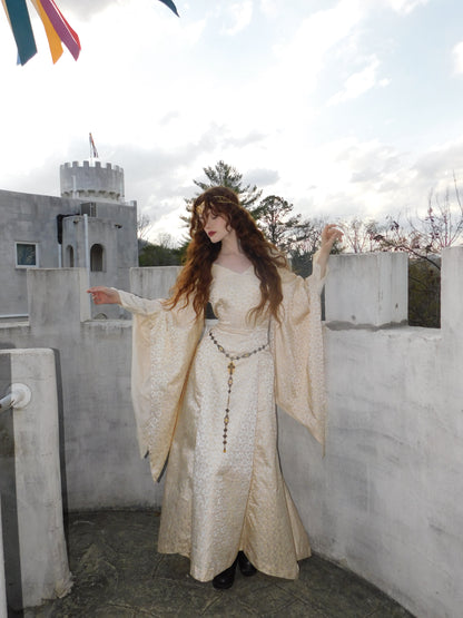 Full-body image of model in embroidered 1930s medieval-style gown with trumpet sleeves and vintage crown, posing on a castle rooftop under festival flags.