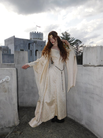 Model wearing medieval-style 1930s vintage gown with long flared sleeves and flowing silhouette, standing on castle balcony at dusk.
