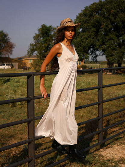 Model in 1970s white maxi slip dress with pastel floral appliqué leaning on ranch fence, styled with cowboy boots and hat