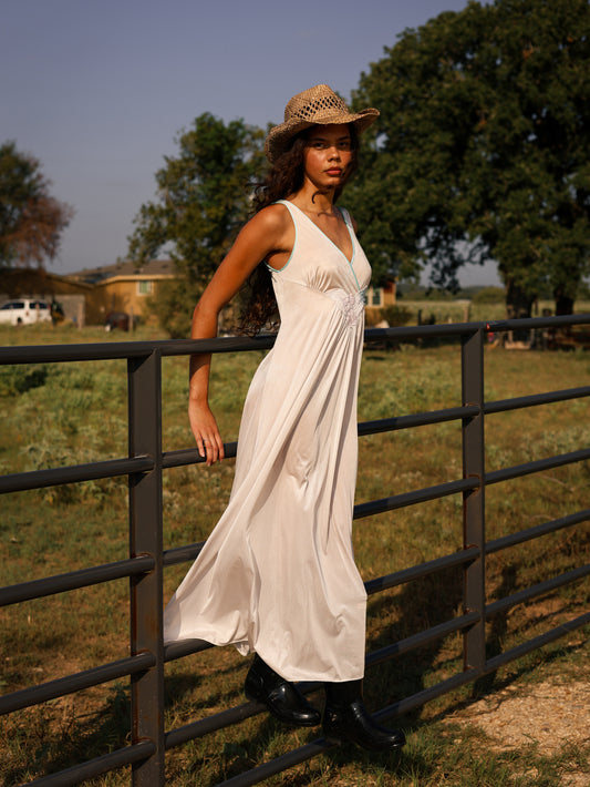 Model in 1970s white maxi slip dress with pastel floral appliqué leaning on ranch fence, styled with cowboy boots and hat