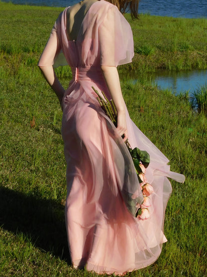 Back view of pink vintage 1960s maxi gown with capelet sleeves and flowing layers – romantic lakeside setting.