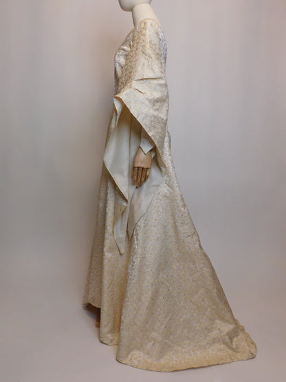 Side profile of a 1930s Spanish silk gown displayed on a mannequin, featuring a floor-length silhouette and exaggerated medieval-inspired sleeves. The ivory silk fabric is embroidered with intricate golden spiral motifs.