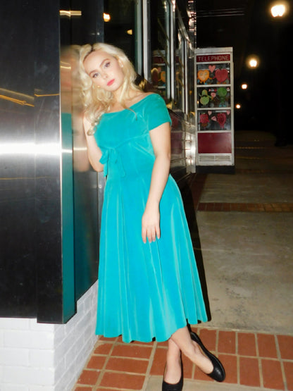 Model in 1950s blue velvet midi dress with bow detail, standing beside a chrome diner at night