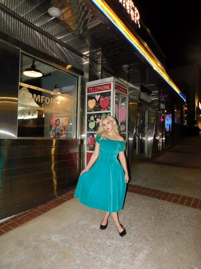 Model wearing a 1950s teal blue velvet midi dress styled with black pumps outside a retro diner at night, next to a vintage telephone booth.