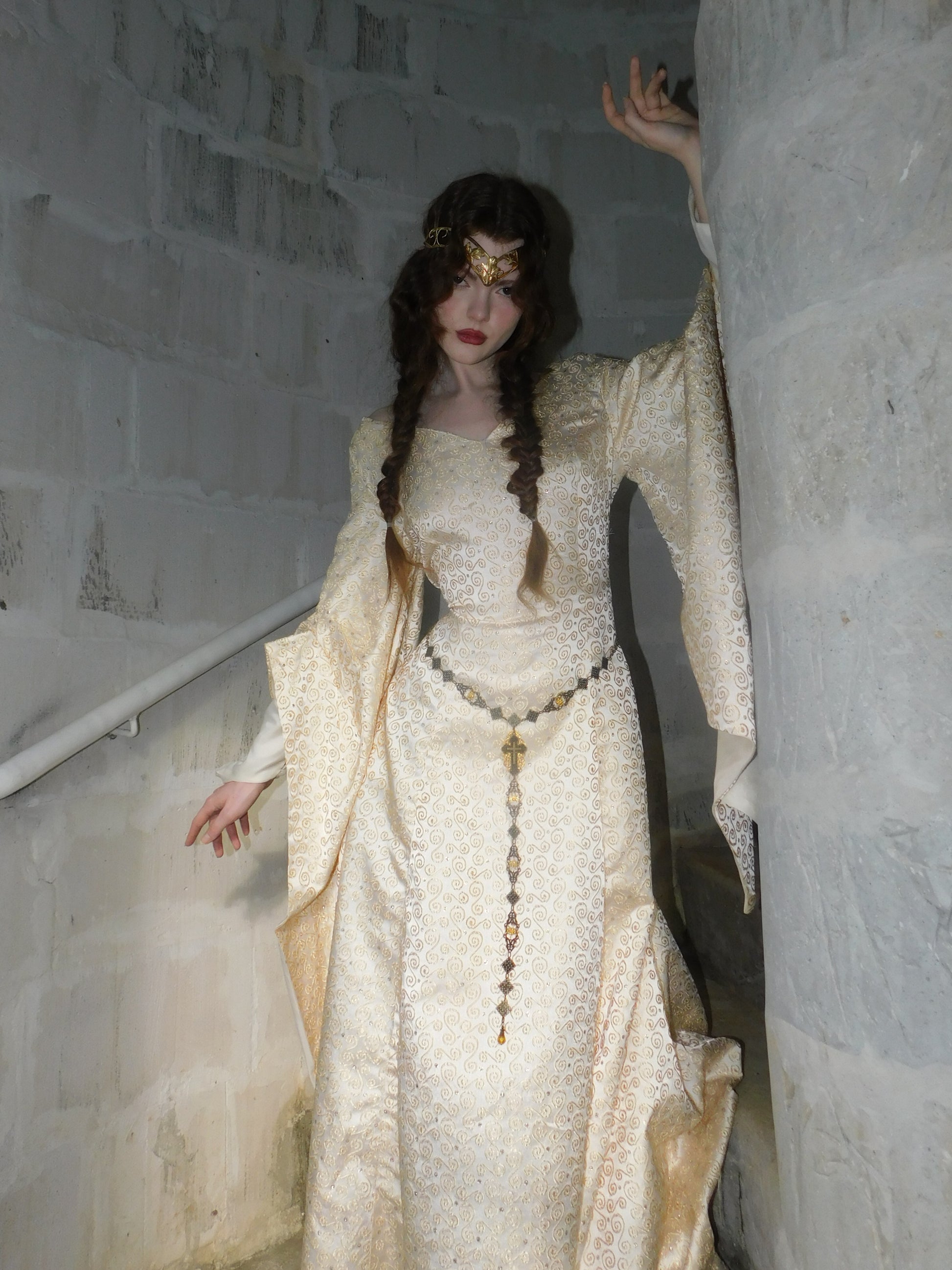 Model wearing a 1930s Spanish silk medieval-style gown with gold embroidery, standing in a stone stairwell with hand gracefully raised.