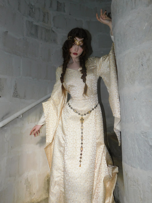 Model wearing a 1930s Spanish silk medieval-style gown with gold embroidery, standing in a stone stairwell with hand gracefully raised.