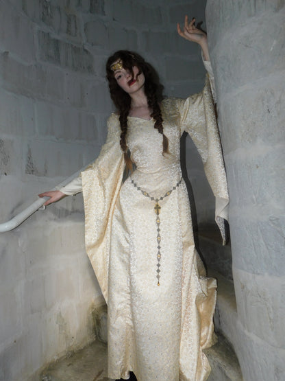Model wearing 1930s vintage Spanish silk gown with gold embroidery, styled with a medieval circlet, raises hand in stone stairwell.