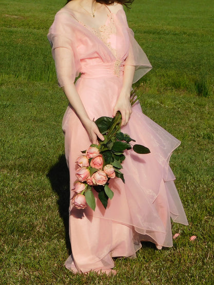 Model in 1960s pink organza maxi dress holding blush roses, standing in a green field – vintage formal gown editorial.