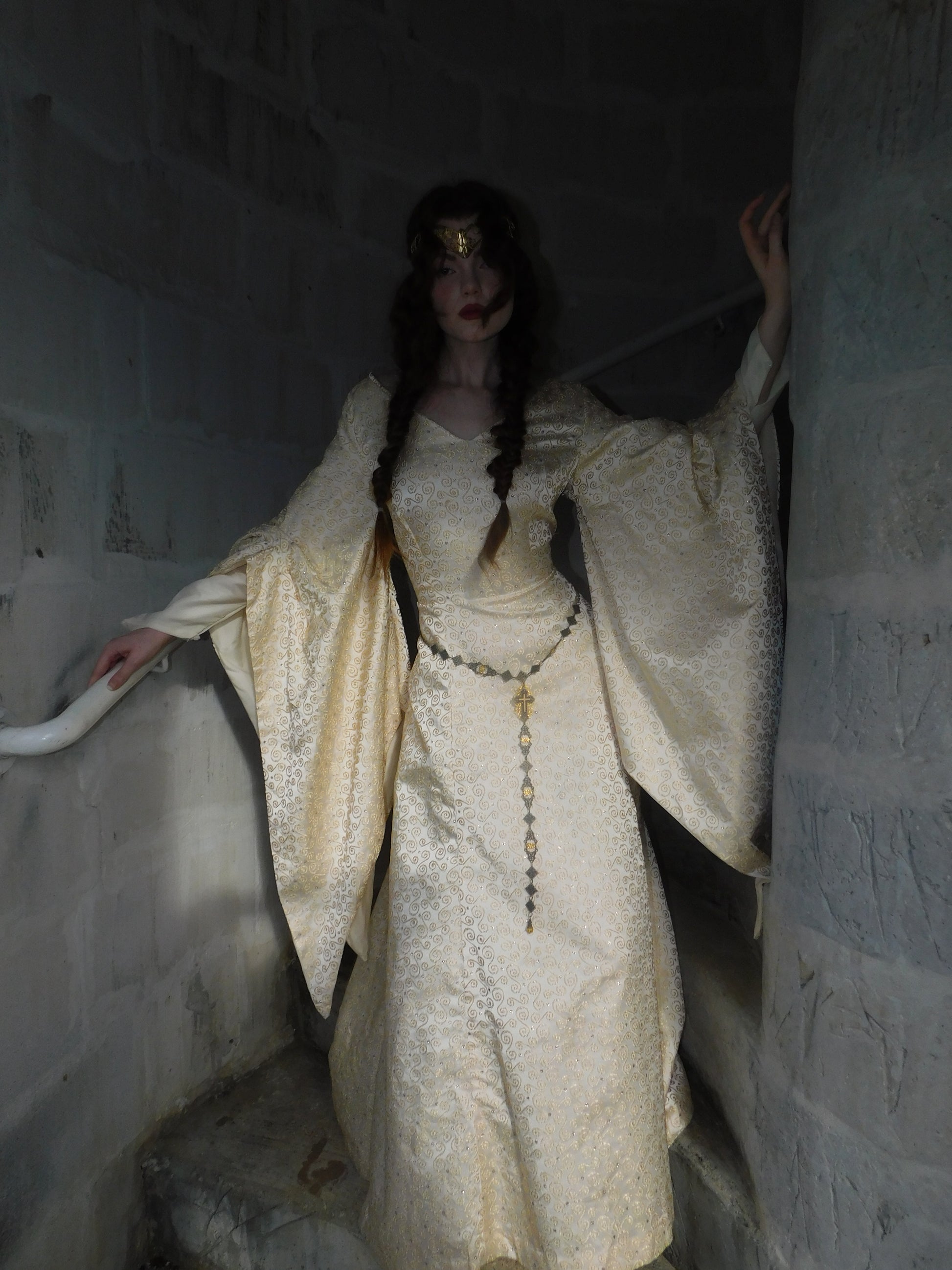 Model in 1930s brocade medieval-inspired gown with flared trumpet sleeves and vintage gold crown, standing in dim castle stairwell.