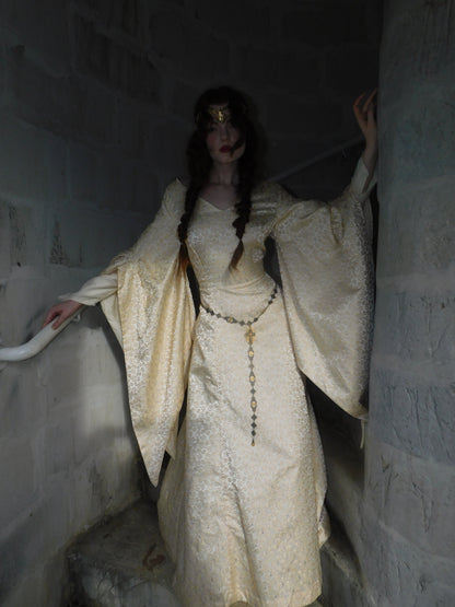 Model in 1930s brocade medieval-inspired gown with flared trumpet sleeves and vintage gold crown, standing in dim castle stairwell.